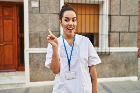 Téléchargez les photos : Jeune femme hispanique portant une carte d'identité de médecin souriant avec une idée ou une question pointant du doigt avec un visage heureux, numéro un - en image libre de droit