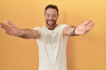 Photo for Middle age man with beard standing over yellow background looking at the camera smiling with open arms for hug. cheerful expression embracing happiness. - Royalty Free Image