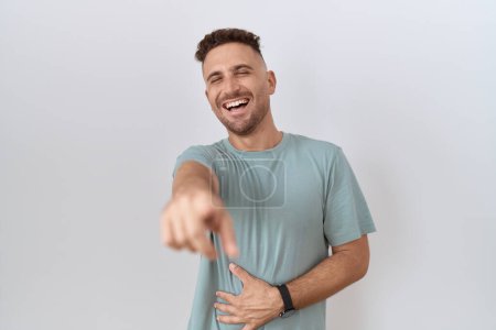 Foto de Hombre hispano con barba de pie sobre fondo blanco riéndose de ti, señalando con el dedo a la cámara con la mano sobre el cuerpo, expresión de vergüenza - Imagen libre de derechos