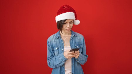 Téléchargez les photos : Jeune femme caucasienne portant un chapeau de Noël en utilisant un smartphone sur fond rouge isolé - en image libre de droit