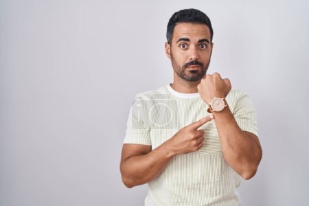 Foto de Hombre hispano con barba de pie sobre un fondo aislado con prisa apuntando a ver el tiempo, la impaciencia, mirando a la cámara con expresión relajada - Imagen libre de derechos