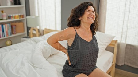 Téléchargez les photos : Moyen âge hispanique femme souffrant de blessures au dos assis sur le lit dans la chambre - en image libre de droit