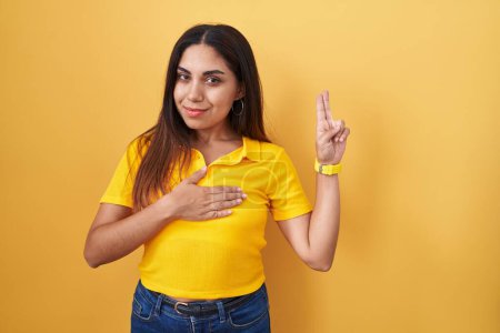 Photo for Young arab woman standing over yellow background smiling swearing with hand on chest and fingers up, making a loyalty promise oath - Royalty Free Image