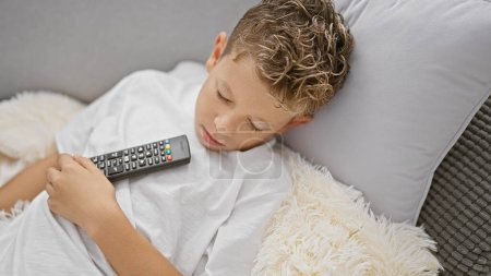 Photo for Blond boy holding tv remote control lying on sofa sleeping at home - Royalty Free Image