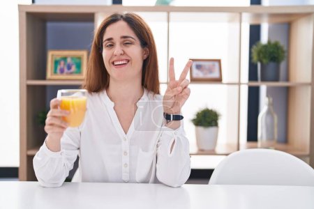 Téléchargez les photos : Femme brune buvant un verre de jus d'orange souriant avec un clin d'œil heureux à la caméra faisant signe de victoire avec les doigts. numéro deux. - en image libre de droit