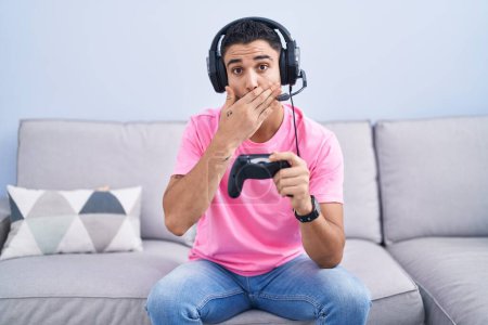 Photo for Hispanic young man playing video game holding controller sitting on the sofa shocked covering mouth with hands for mistake. secret concept. - Royalty Free Image