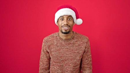 Photo for African american man surprise expression wearing christmas hat over isolated red background - Royalty Free Image