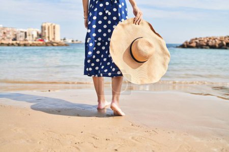 Foto de Joven mujer rubia turista sosteniendo sombrero de verano caminando en la playa - Imagen libre de derechos
