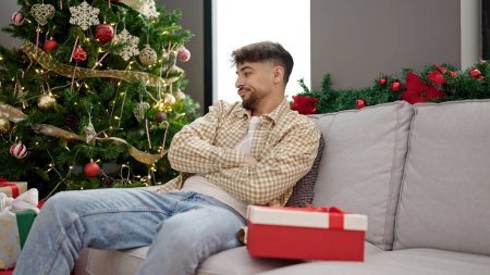 Photo for Young arab man unpacking gift looking upset sitting by christmas tree at home - Royalty Free Image