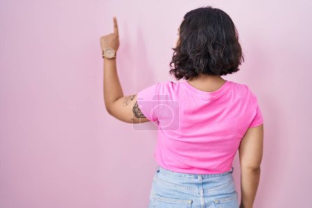 Foto de Young hispanic woman standing over pink background posing backwards pointing ahead with finger hand - Imagen libre de derechos