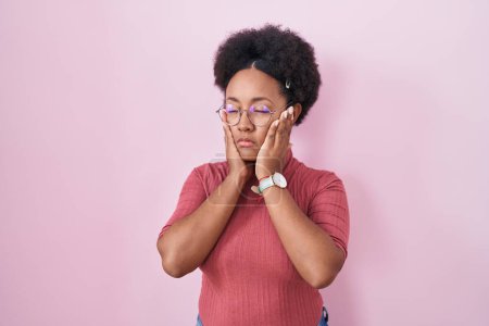 Téléchargez les photos : Belle femme africaine avec les cheveux bouclés debout sur fond rose mains fatiguées couvrant le visage, la dépression et la tristesse, contrarié et irrité par le problème - en image libre de droit