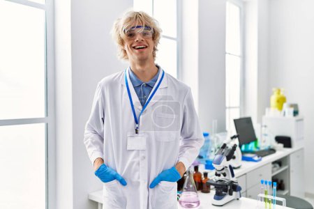 Foto de Joven científico hombre rubio sonriendo confiado de pie en el laboratorio - Imagen libre de derechos