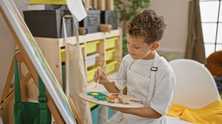 Téléchargez les photos : Adorable garçon artiste blond assis avec confiance sur une chaise, dessinant dans un studio d'art intérieur, incarnant la créativité - en image libre de droit