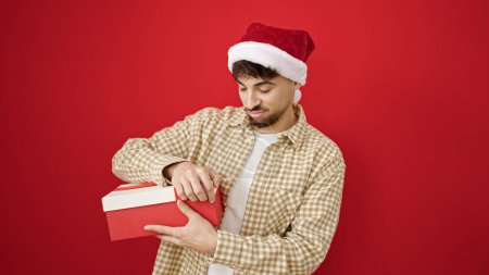 Téléchargez les photos : Jeune homme arabe portant chapeau de Noël déballage cadeau regardant bouleversé sur fond rouge isolé - en image libre de droit