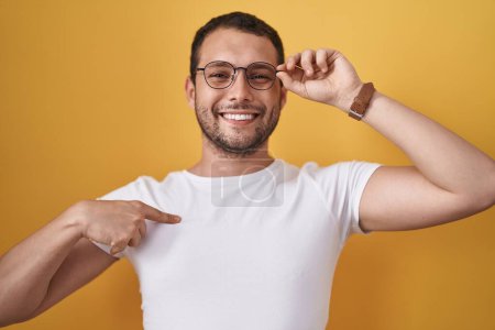 Foto de Hombre hispano sosteniendo gafas señalando con el dedo a uno mismo sonriendo feliz y orgulloso - Imagen libre de derechos