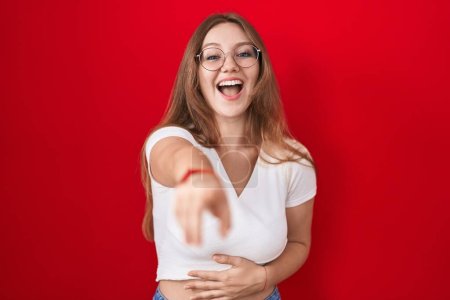 Foto de Joven mujer caucásica de pie sobre fondo rojo riéndose de ti, señalando con el dedo a la cámara con la mano sobre el cuerpo, expresión de vergüenza - Imagen libre de derechos