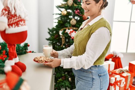 Photo for Young beautiful hispanic woman holding cookies and glass of milk standing by christmas tree at home - Royalty Free Image