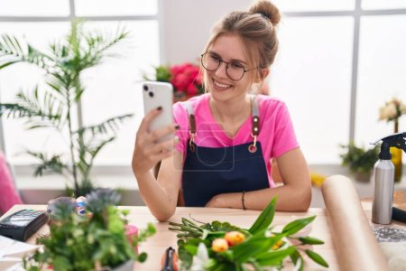 Foto de Joven florista mujer rubia sonriendo confiado hacer selfie por teléfono inteligente en la tienda de flores - Imagen libre de derechos
