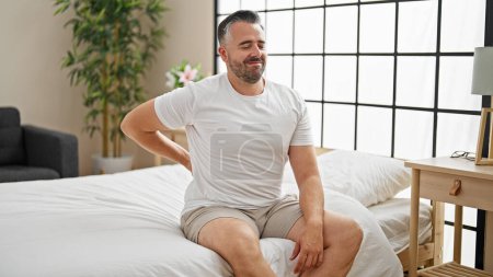 Téléchargez les photos : Homme aux cheveux gris souffrant d'une blessure au dos assis sur le lit dans la chambre - en image libre de droit