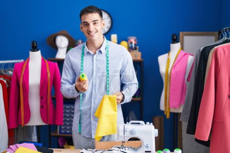 Téléchargez les photos : Jeune homme hispanique tailleur souriant confiance tenant tissu et fil au studio de couture - en image libre de droit