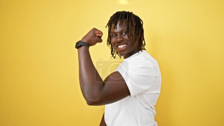 Téléchargez les photos : Afro-Américain souriant confiant faire un geste fort avec le bras sur fond jaune isolé - en image libre de droit