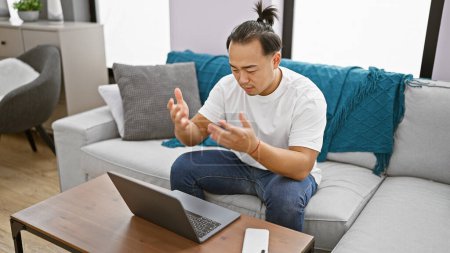 Photo for Handsome young chinese man, focused and concentrated, working online using laptop while sitting on sofa indoors. delving deep into the world of internet technology at his apartment's living room. - Royalty Free Image
