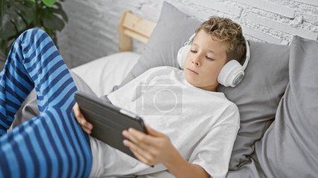 Entzückender, kleiner, blonder Junge, der ernsthaft in sich vertieft ist, Video auf dem Touchpad anschaut, während er sich im Bett entspannt, in der gemütlichen Umgebung seines Schlafzimmers drinnen.