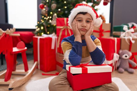 Foto de Adorable niño hispano sosteniendo regalo de Navidad sentado en el suelo con expresión infeliz en casa - Imagen libre de derechos