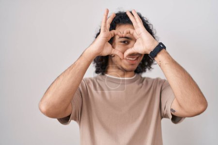 Photo for Hispanic man with curly hair standing over white background doing heart shape with hand and fingers smiling looking through sign - Royalty Free Image