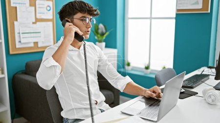 Téléchargez les photos : Jeune adolescent hispanique parler affaires au téléphone, travailler dur avec ordinateur portable au bureau - en image libre de droit