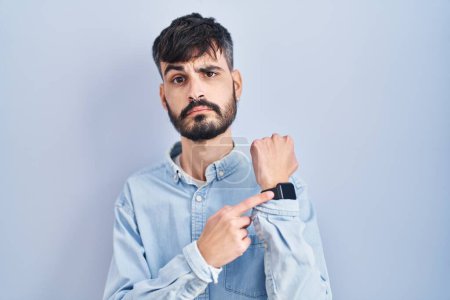 Téléchargez les photos : Jeune homme hispanique avec la barbe debout sur fond bleu pressé pointant vers regarder le temps, l'impatience, regardant la caméra avec une expression détendue - en image libre de droit