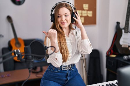 Téléchargez les photos : Jeune femme caucasienne enregistrement chanson au studio de musique pointant pouce vers le côté souriant heureux avec bouche ouverte - en image libre de droit