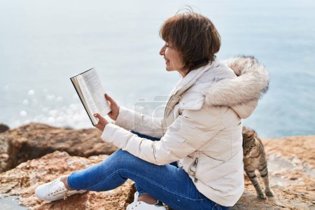 Téléchargez les photos : Middle age woman reading book sitting on the rock at seaside - en image libre de droit