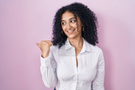 Foto de Hispanic woman with curly hair standing over pink background smiling with happy face looking and pointing to the side with thumb up. - Imagen libre de derechos