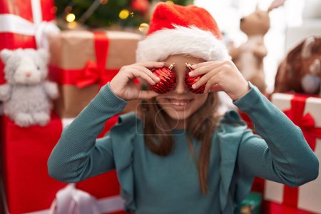 Téléchargez les photos : Adorable fille hispanique tenant boules décoration sur les yeux debout près de l'arbre de Noël à la maison - en image libre de droit