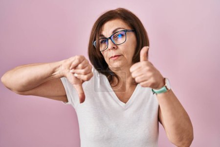 Foto de Middle age hispanic woman standing over pink background doing thumbs up and down, disagreement and agreement expression. crazy conflict - Imagen libre de derechos