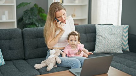 Foto de Madre e hija sentadas en el sofá hablando en el teléfono inteligente usando el ordenador portátil estresado en casa - Imagen libre de derechos