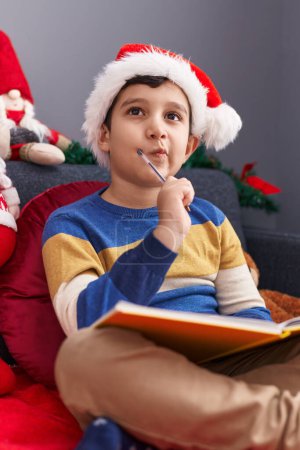Photo for Adorable hispanic boy writing on notebook sitting on sofa by christmas decoration at home - Royalty Free Image