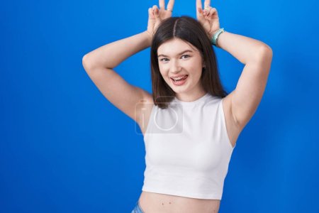 Téléchargez les photos : Jeune femme caucasienne debout sur fond bleu posant drôle et fou avec les doigts sur la tête comme des oreilles de lapin, souriant gai - en image libre de droit