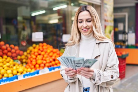 Photo for Young blonde woman smiling confident counting dollars at fruit store - Royalty Free Image