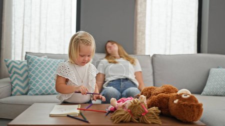 Photo for Stressed mother relaxing on sofa at home while little caucasian daughter seriously engages in drawing on notebook indoors - Royalty Free Image