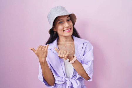 Photo for Young hispanic woman standing over pink background wearing hat pointing to the back behind with hand and thumbs up, smiling confident - Royalty Free Image