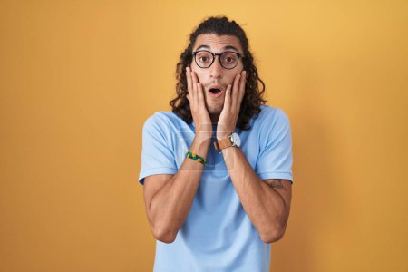 Photo for Young hispanic man standing over yellow background afraid and shocked, surprise and amazed expression with hands on face - Royalty Free Image