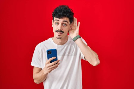 Téléchargez les photos : Homme hispanique utilisant smartphone sur fond rouge souriant avec la main sur l'oreille en écoutant une rumeur ou des commérages. concept de surdité. - en image libre de droit