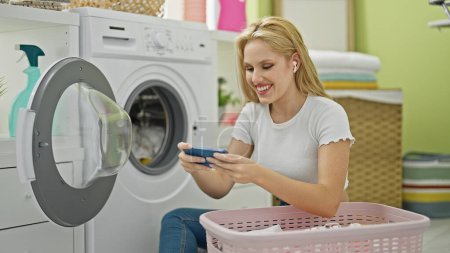 Téléchargez les photos : Jeune femme blonde jouant jeu vidéo lavage des vêtements à la buanderie - en image libre de droit