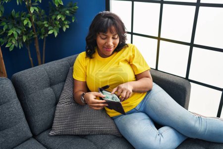 Photo for Young beautiful latin woman holding dollars on wallet sitting on sofa at home - Royalty Free Image