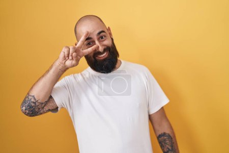 Téléchargez les photos : Jeune homme hispanique avec barbe et tatouages debout sur fond jaune faisant symbole de paix avec les doigts sur le visage, souriant joyeux montrant la victoire - en image libre de droit