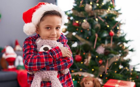 Foto de Adorable niño hispano abrazando oso de peluche de pie junto al árbol de Navidad en casa - Imagen libre de derechos