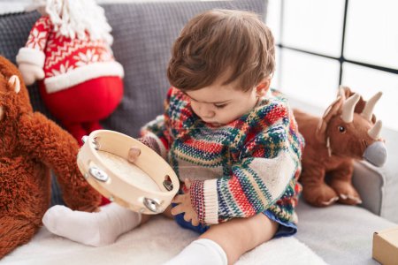 Photo for Adorable hispanic toddler playing tambourine sitting on sofa by christmas tree at home - Royalty Free Image