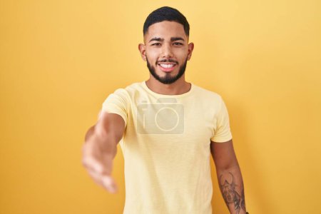 Photo for Young hispanic man standing over yellow background smiling friendly offering handshake as greeting and welcoming. successful business. - Royalty Free Image